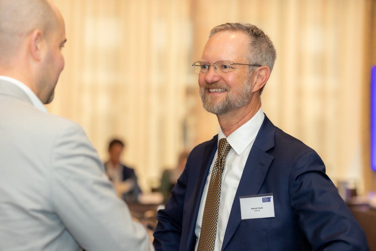 Portrait of David Clark, Chief Sustainability Officer at Amcor, wearing glasses and a dark suit jacket over a light blue shirt. He has short, gray hair and a beard, smiling slightly against a plain gray background.