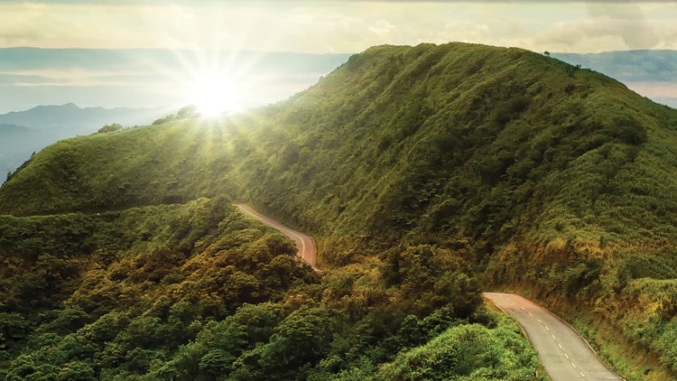 A winding road cutting through lush green hills, with the sun shining brightly over the horizon, casting a warm glow over the landscape.