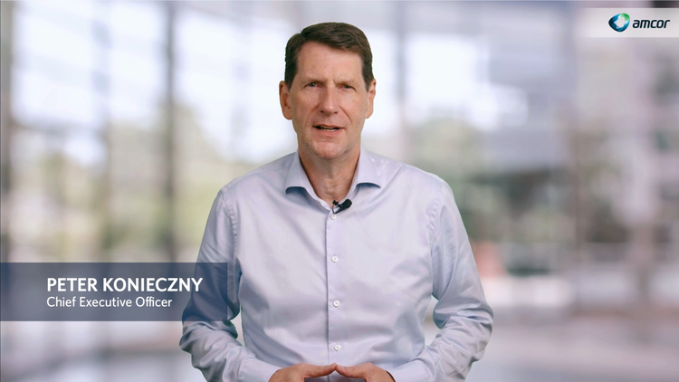 Peter Konieczny, Chief Executive Officer of Amcor, standing in front of a blurred office background. He is wearing a light blue shirt with a microphone clipped on, speaking directly to the camera. The Amcor logo is visible in the top right corner.